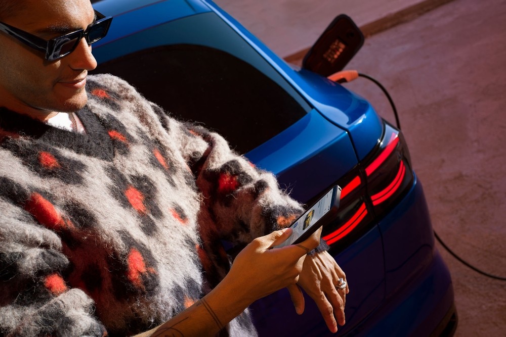 Persoon met zonnebril gebruikt smartphone naast een blauwe Ford elektrische auto die oplaadt, toont de FordApp.