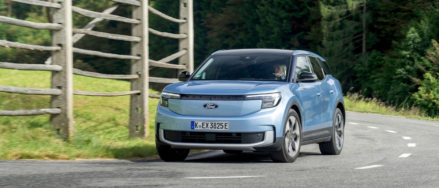 Brede opname van de volledig elektrische Ford Explorer (blauw) op de open weg voor een houten hek.