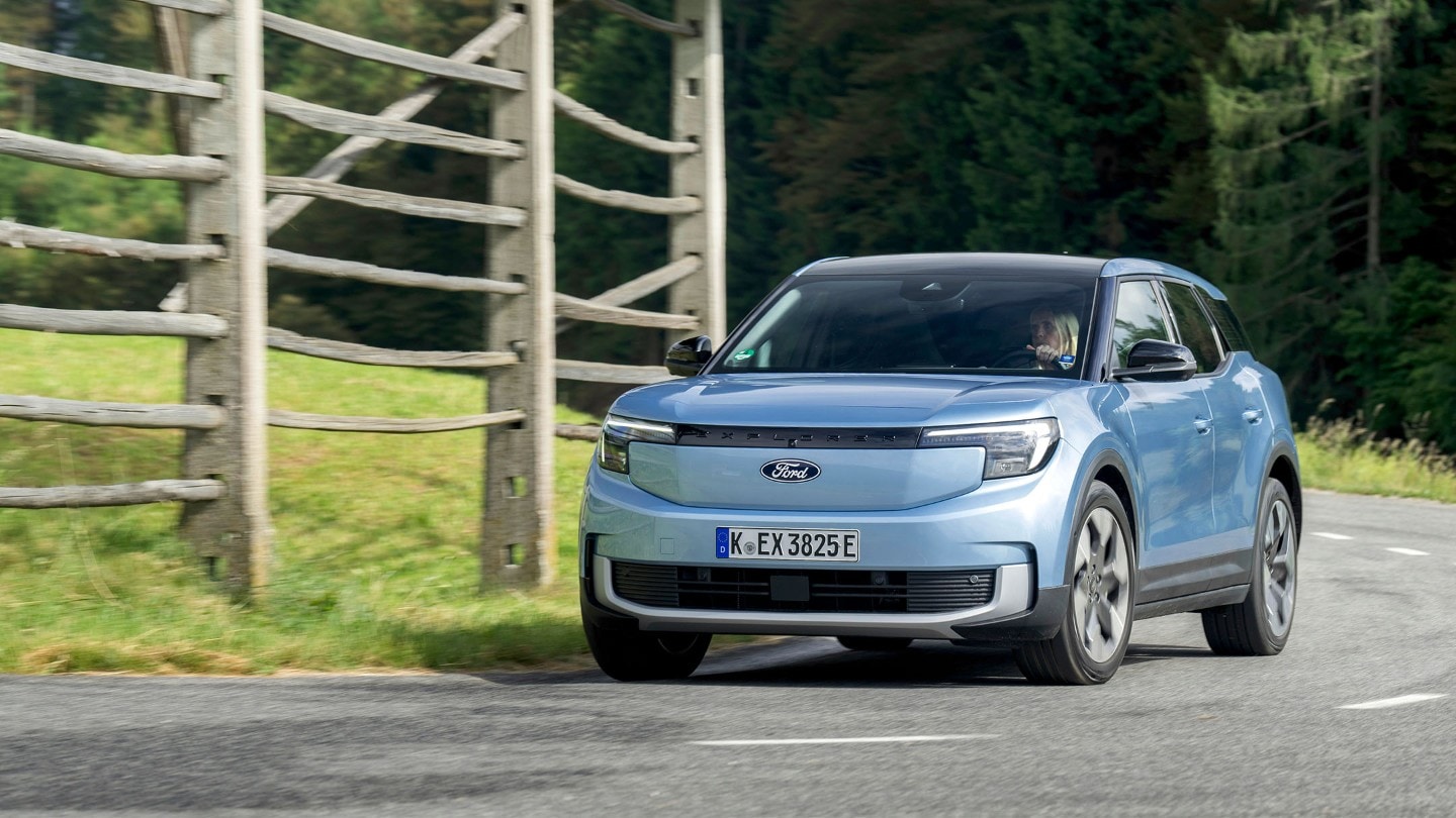 Brede opname van de volledig elektrische Ford Explorer (blauw) op de open weg voor een houten hek.
