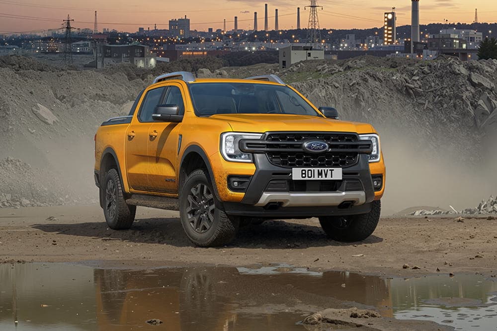 Een robuuste gele Ford Ranger pick-up op een modderig bouwterrein tijdens zonsondergang, met op de achtergrond een industriële skyline.