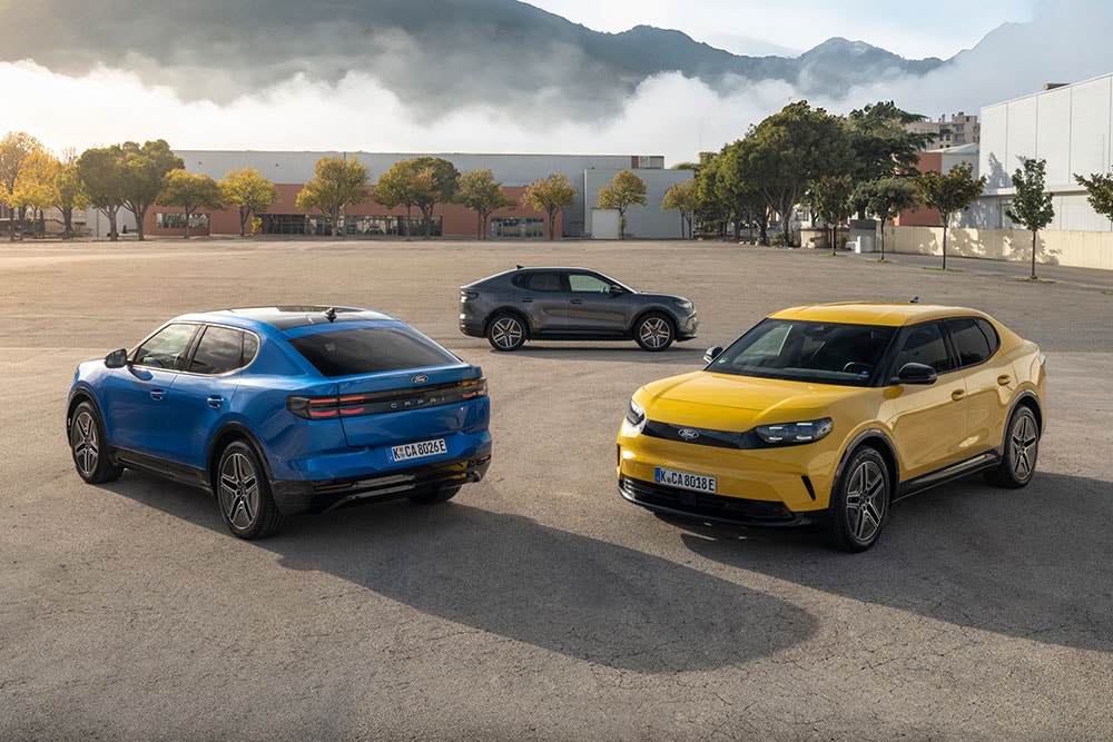 Een blauwe en gele Ford Capri staan geparkeerd op een buitenterrein, met een donkergrijze Capri op de achtergrond.