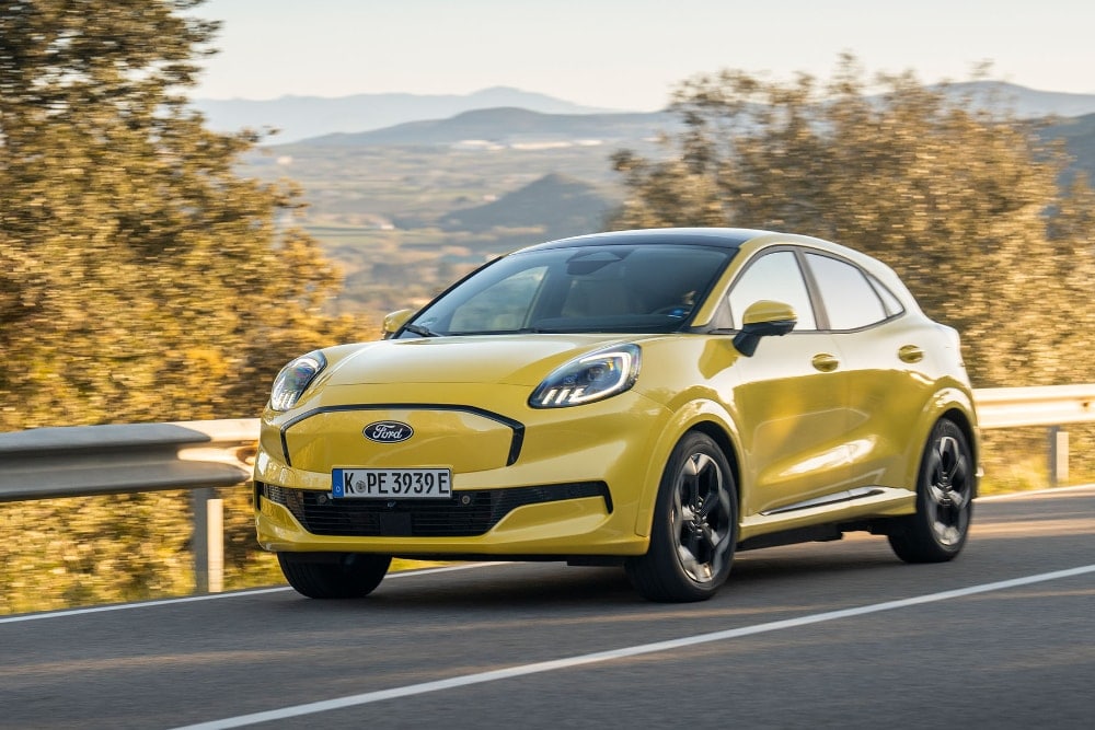 Een gele Ford Puma Gen-E rijdt op een weg met bomen en bergen op de achtergrond.
