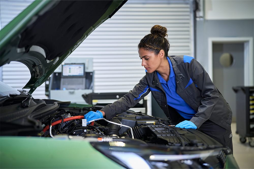 Een technicus met blauwe handschoenen werkt aan de motor van een groene auto in een werkplaats.