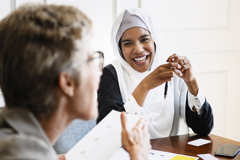 Een vrouw met een witte hijab lacht tijdens een vergadering, met een pen in haar hand. Papieren en een telefoon liggen op tafel. 