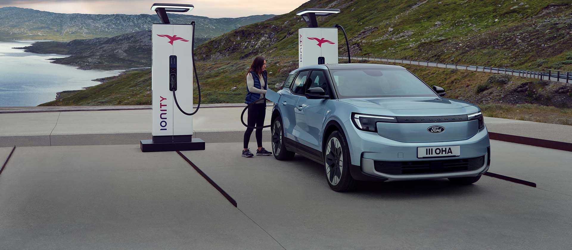 Een vrouw sluit een blauwe, volledig elektrische Ford Explorer aan op een openbaar laadpunt met een kabel. De auto staat geparkeerd bij bergen en water.