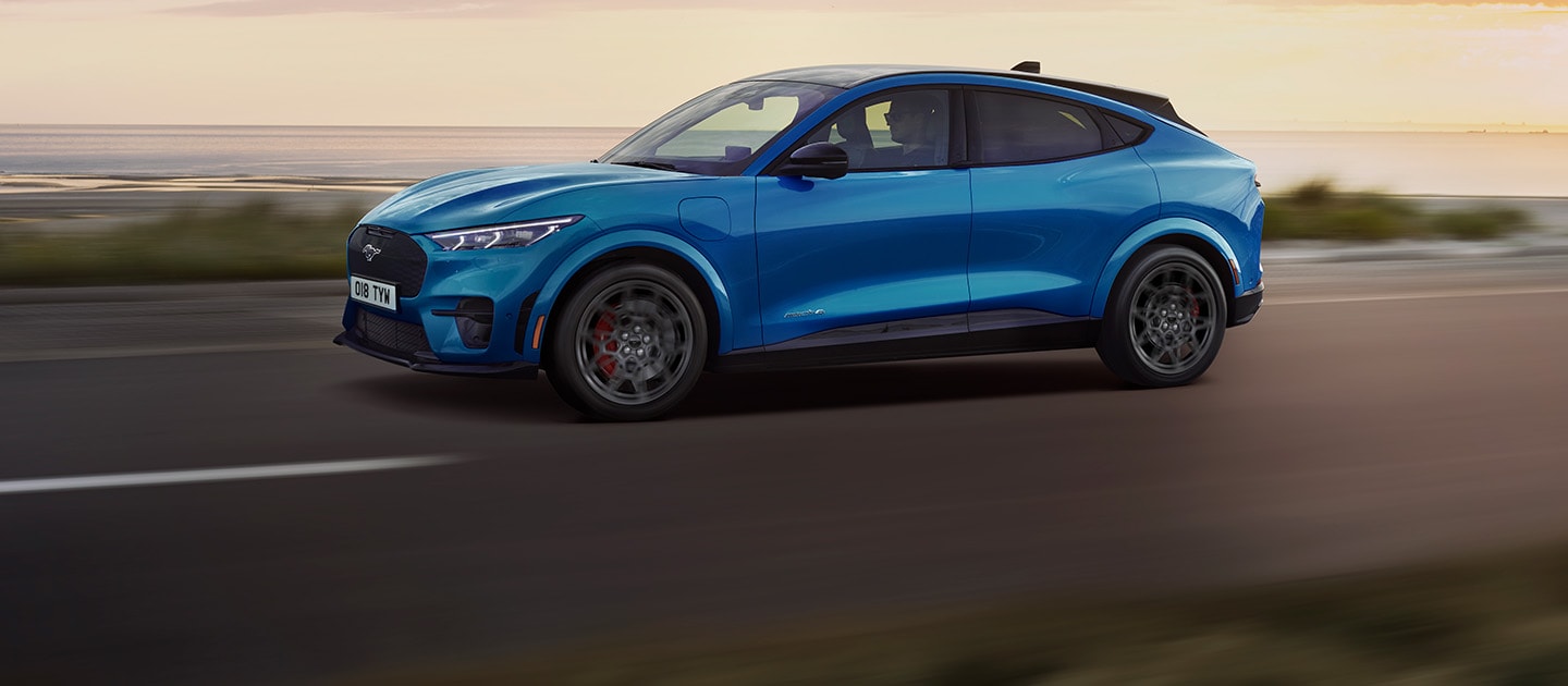 Een blauwe elektrische Ford Mustang Mach-E rijdt over een kustweg bij zonsondergang.