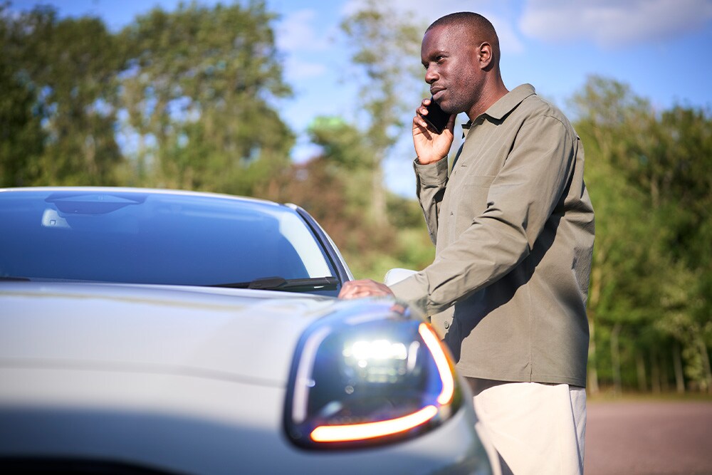 A person on a phone call stands next to a light-coloured car outdoors.