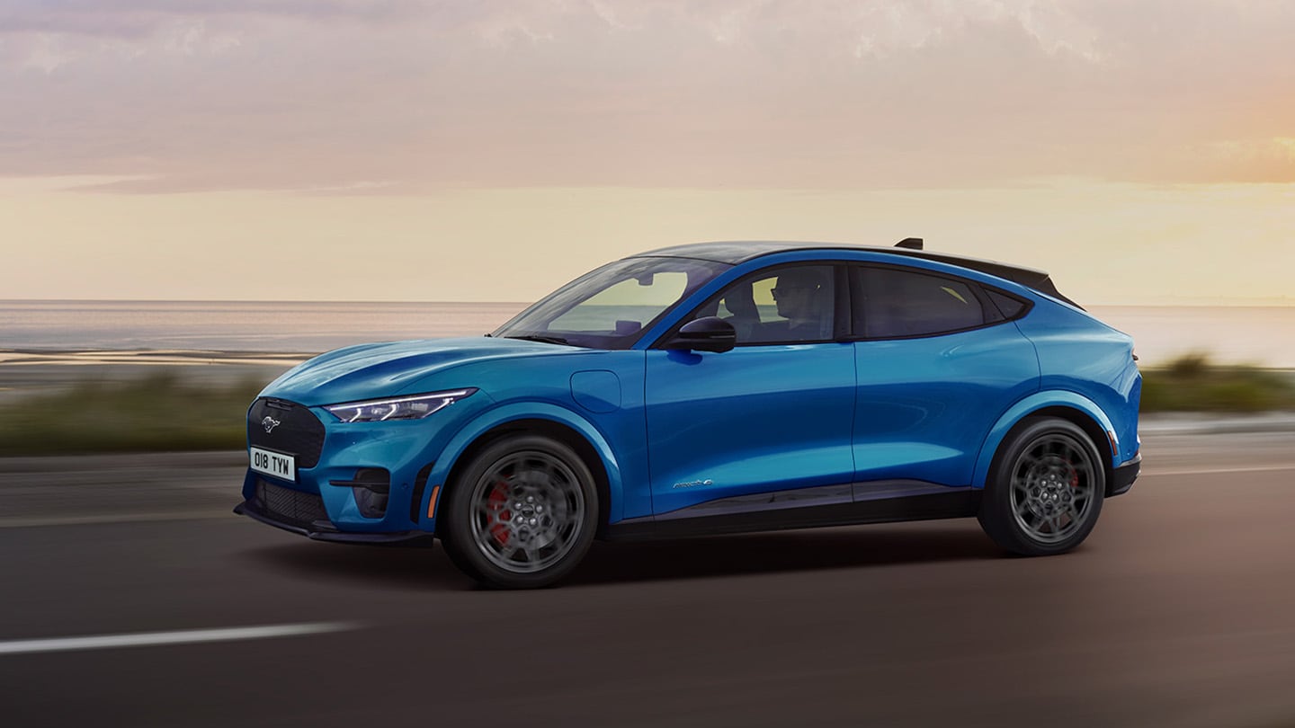 Een zijaanzicht van een blauwe elektrische Ford Mustang Mach-E die in de schemering over een kustweg rijdt.