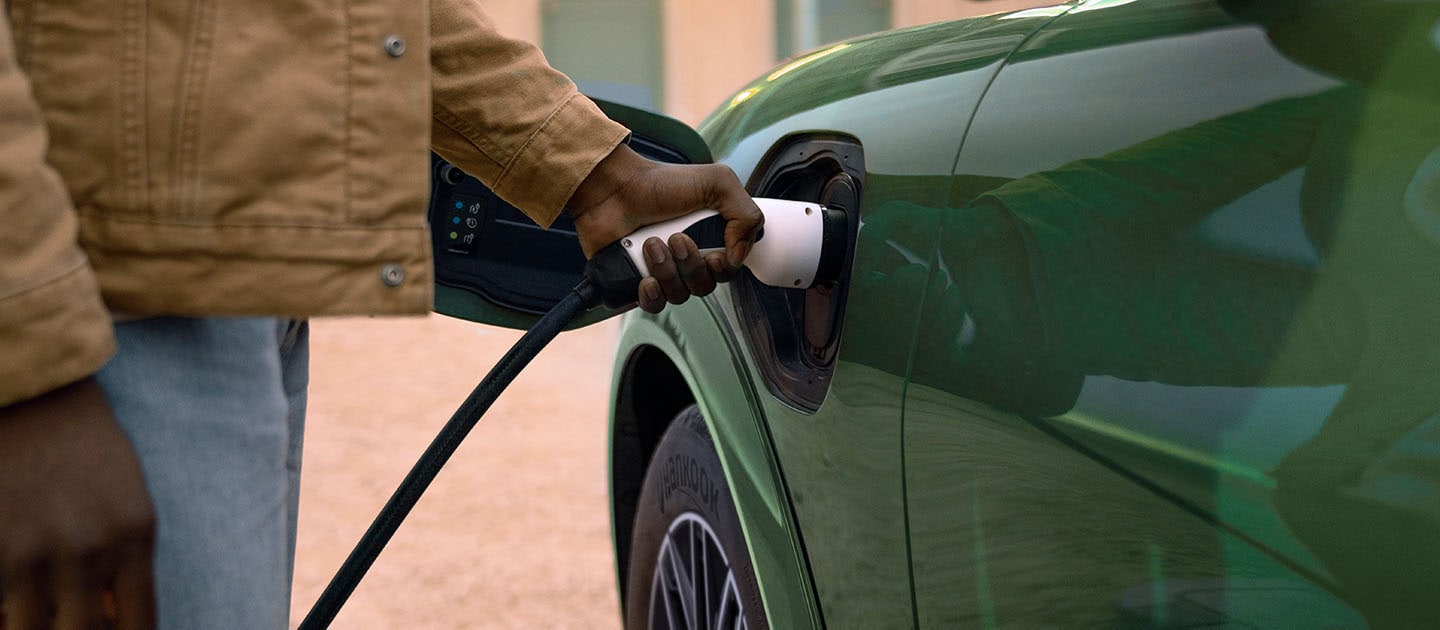 Iemand in een crèmekleurige jas sluit een elektrische oplaadkabel aan op een groene, elektrische Ford Mustang Mach-E.
