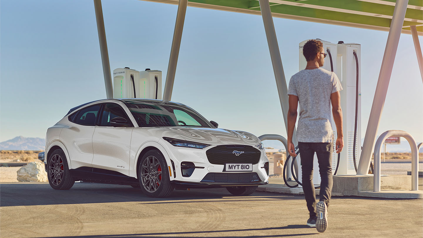 Een witte Ford Mustang Mach-E geparkeerd bij een openbaar laadpunt. Een man in een wit T-shirt loopt naar de auto.