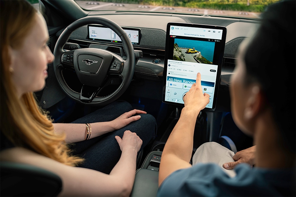 Close-up van het dashboard van een Mustang Mach-E, waarbij een bestuurder naar het touchscreen wijst.