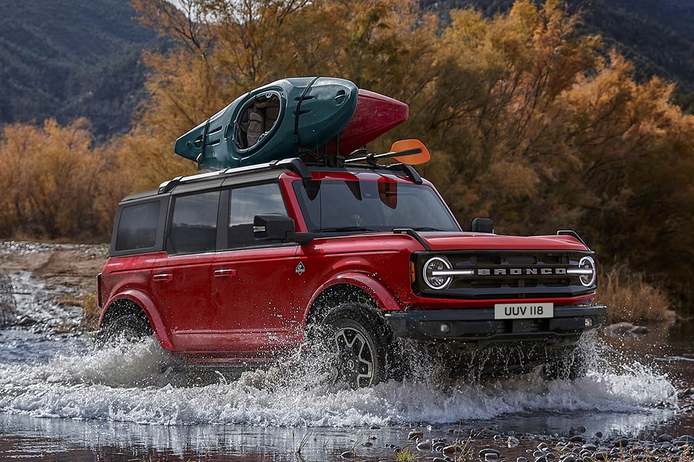 Side view of a red Ford Bronco driving through shallow water in a mountain setting.
