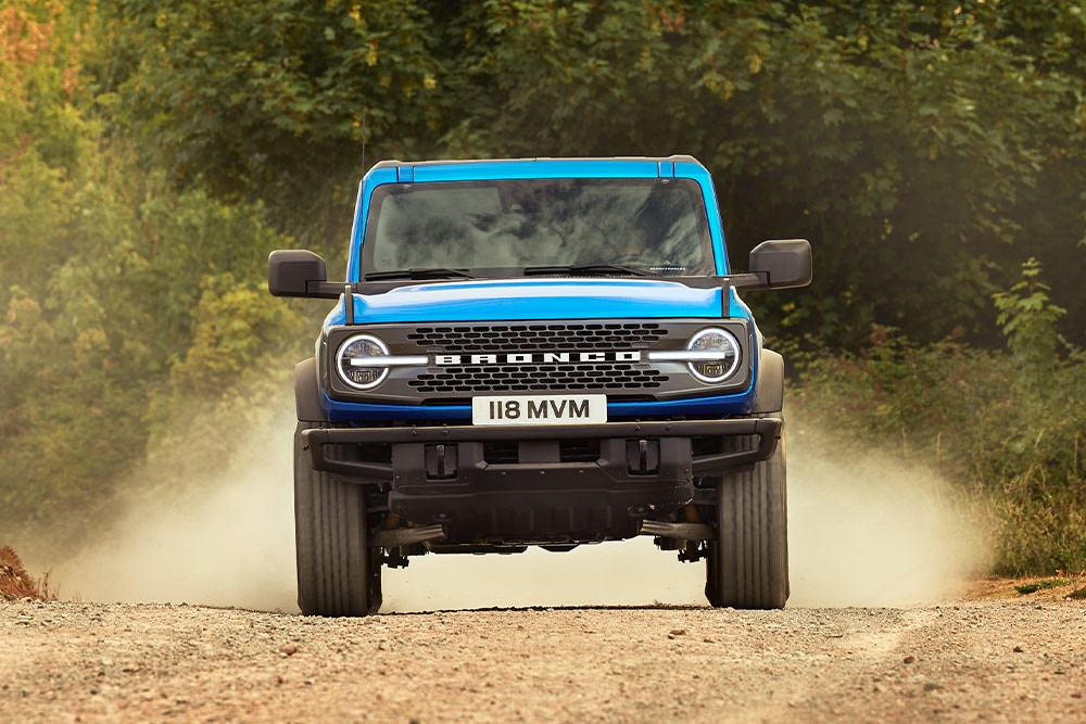 Front view of a blue Ford Bronco driving on a dirt road. 