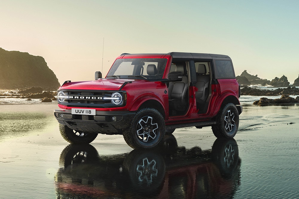 Side view of a red Ford Bronco, with doors removed, parked on a wet sandy shore.