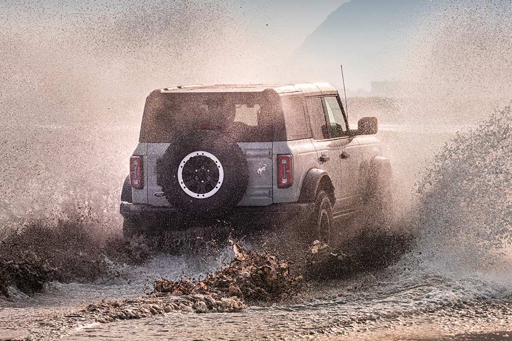 A rear view of a silver Ford Bronco driving through a spray of water on a beach.