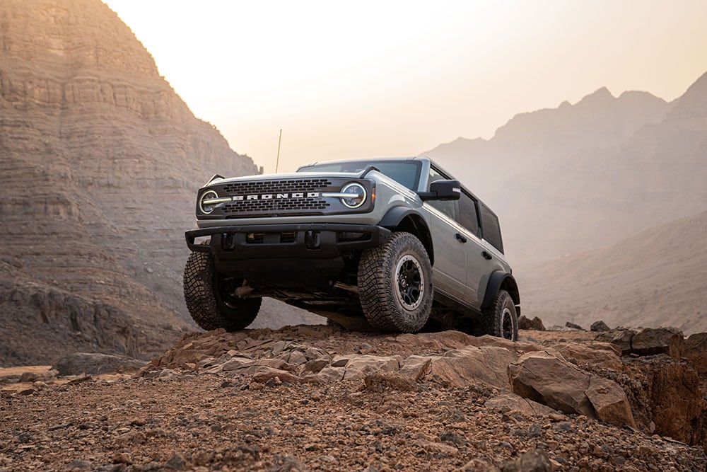 A grey Ford Bronco parked atop a rocky path in a mountain setting.