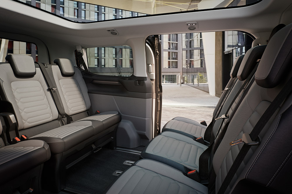 Interieurbeeld van de Ford Tourneo Custom, met meerdere rijen grijze passagiersstoelen en panoramisch dak.