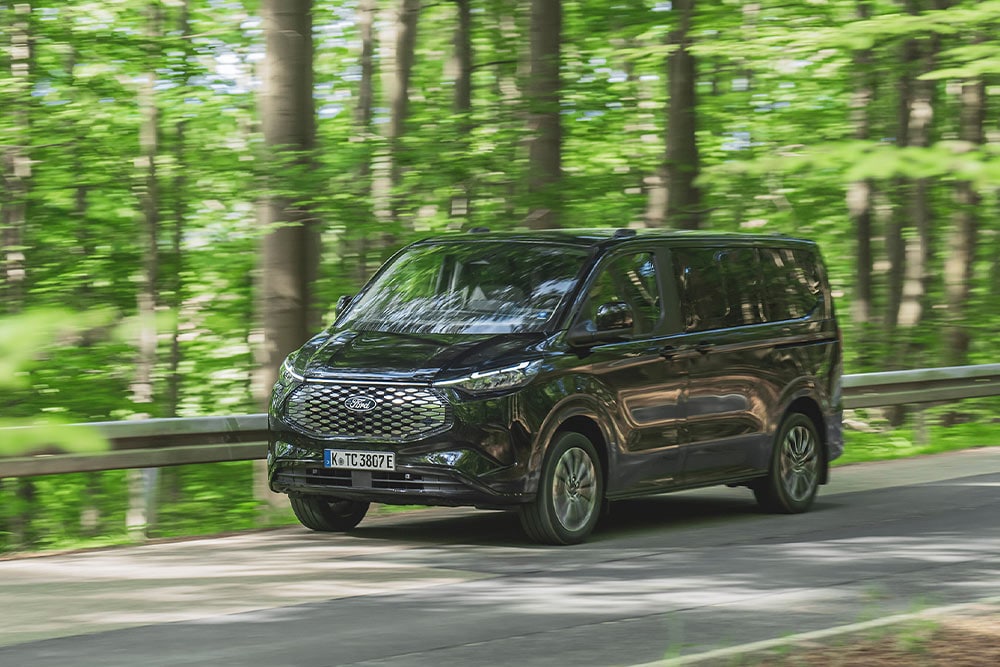 Een zwarte Ford E-Tourneo Custom rijdt over een met bomen omzoomde weg.