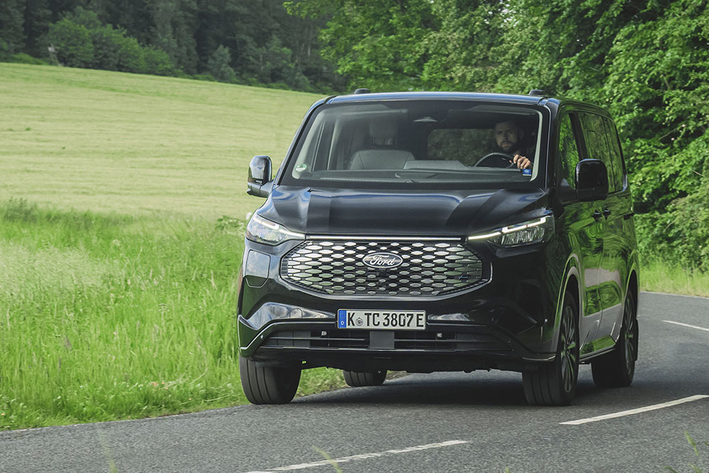 Een zwarte Ford E-Tourneo Custom rijdt over een landweg.