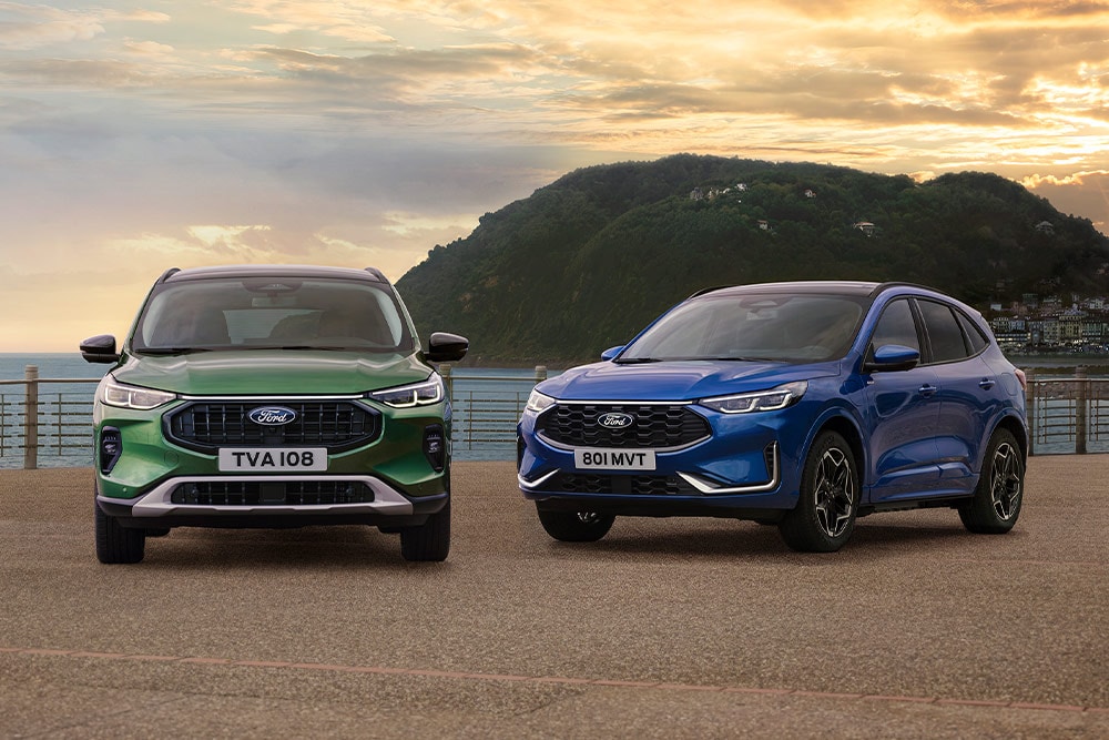 Een groene Ford Kuga Active en een blauwe Ford Kuga ST Line naast elkaar geparkeerd op een parkeerplaats aan de kust. 