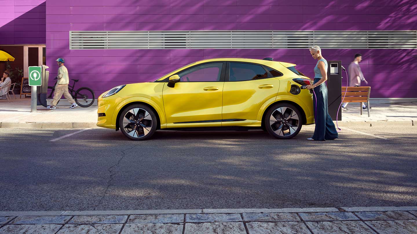 Zijaanzicht van een gele elektrische Ford Puma Gen-E® die wordt aangesloten op een openbare laadpaal op een parkeerplaats.