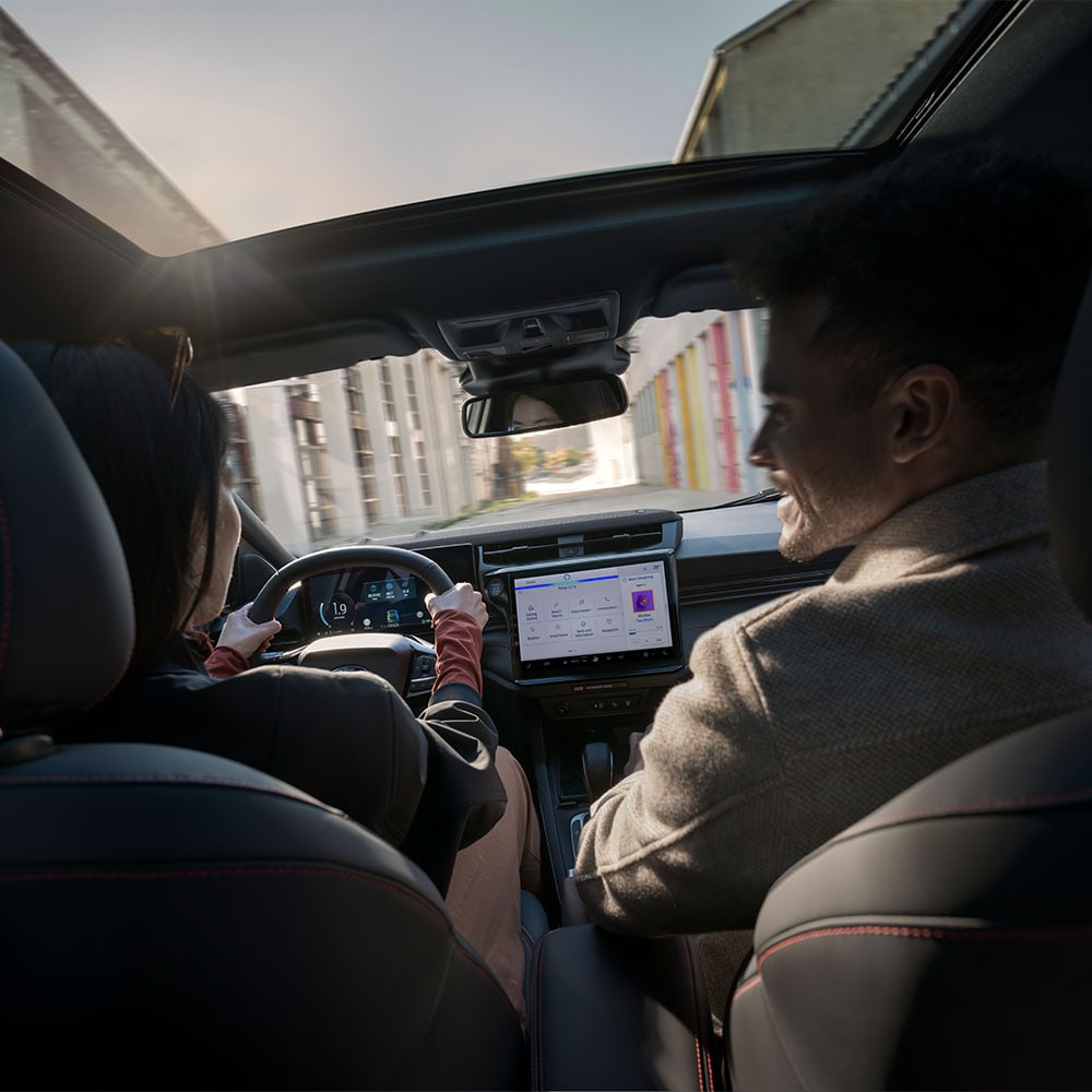 Een chauffeur en passagier kletsen terwijl ze door een zonnig woonlandschap rijden.