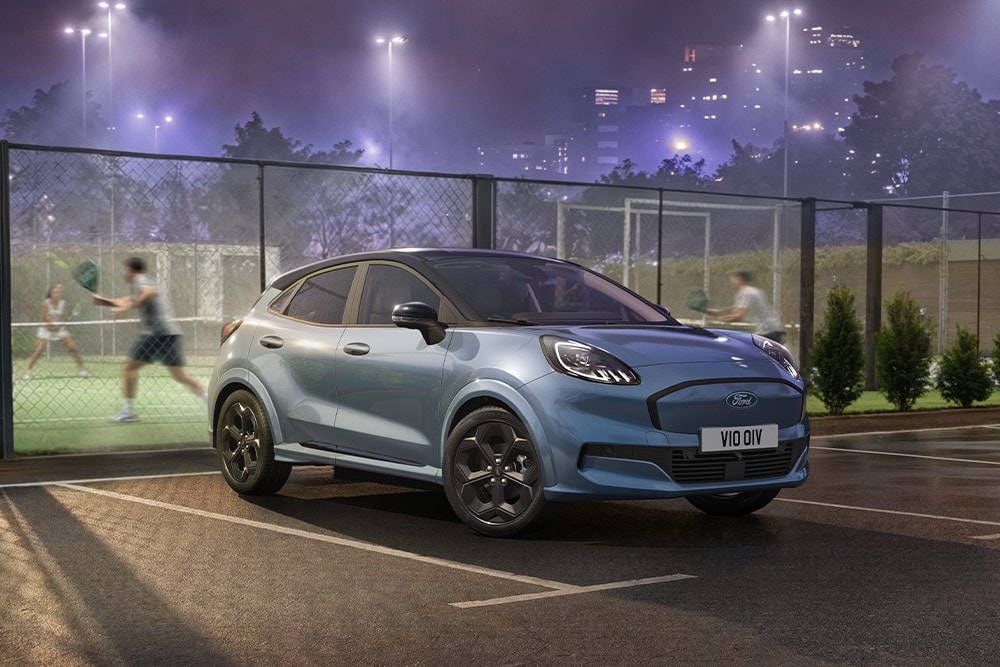 Een blauwe Ford Puma Gen-E die geparkeerd staat naast een tennisbaan, met stadslichten op de achtergrond.