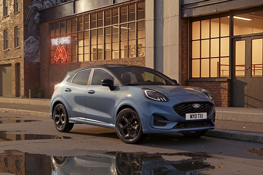 Een blauwe Ford Puma die geparkeerd staat op een stedelijke straat bij zonsondergang, met plassen waarin de gebouwen weerspiegeld zijn.