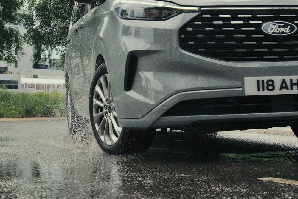 Het voorwiel van een zilveren Ford Tourneo Custom spettert door een plas.