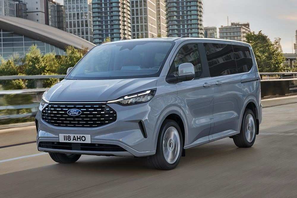Een zilveren Ford Tourneo Custom rijdt op een stadsweg.