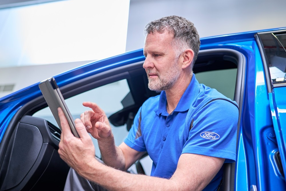 Een monteur zit op de drempel van een blauwe Ford en gebruikt een tablet voor een diagnose.