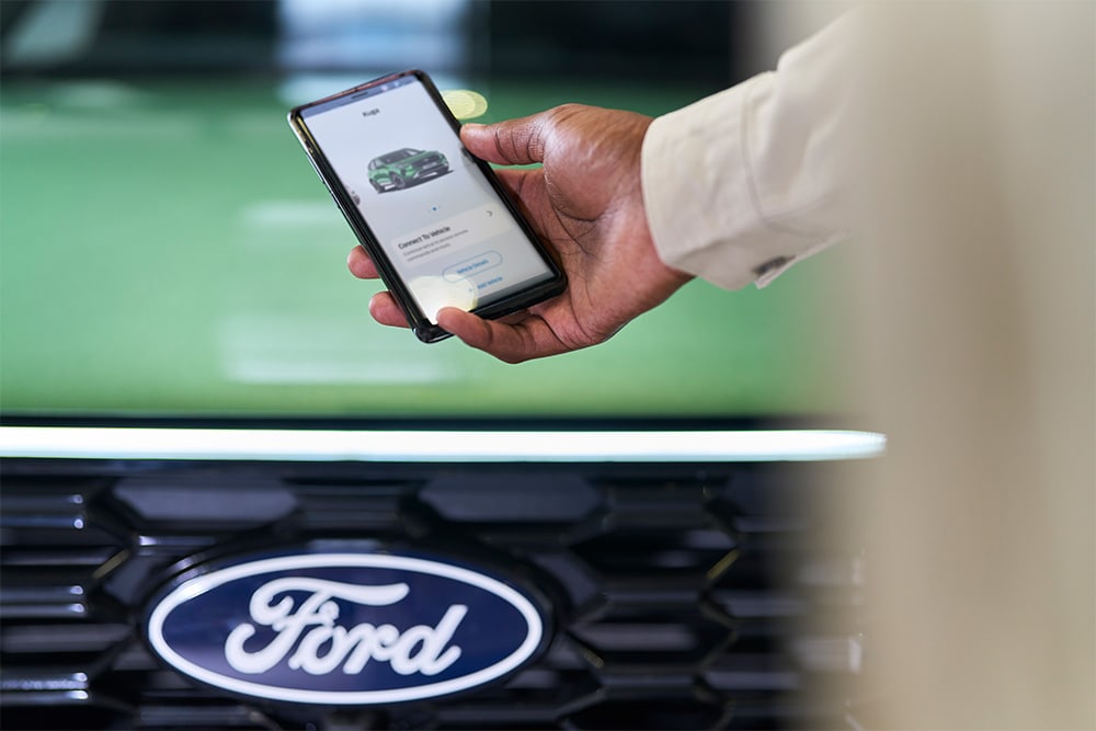 A person's hand holding a smartphone displaying the FordApp with a Ford Kuga, above a Ford grille.