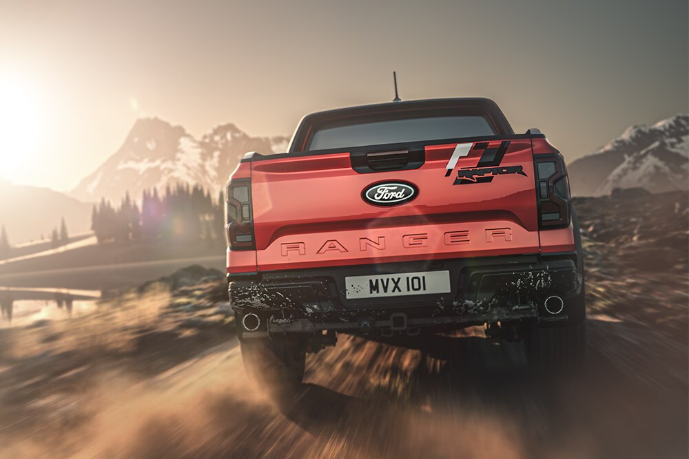 Achteraanzicht van een Ford Ranger Raptor die over een onverharde weg rijdt, met bergen op de achtergrond.