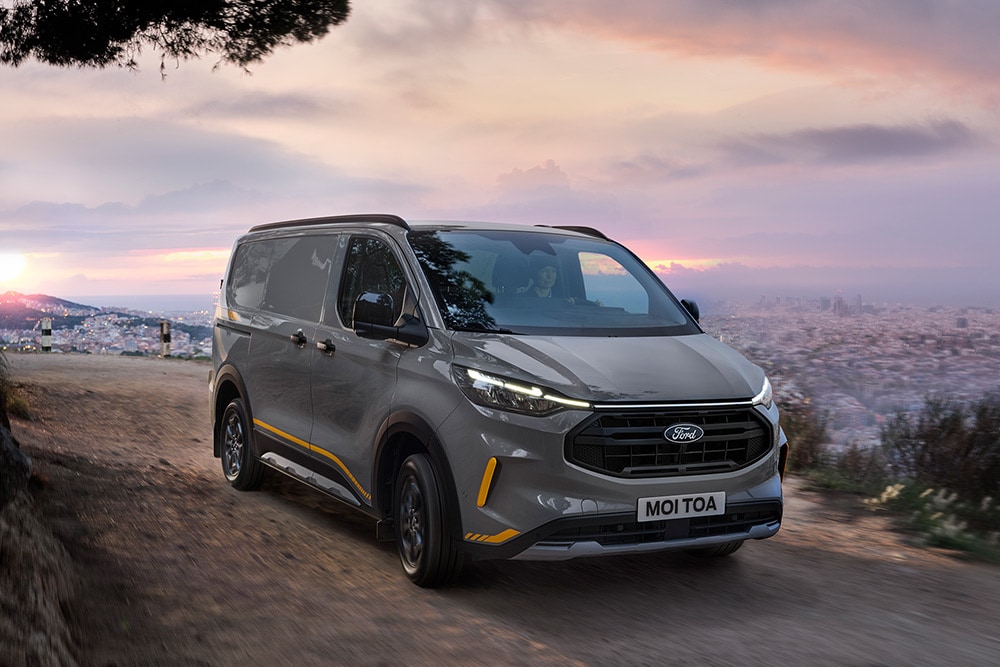 Een grijze Ford Transit Custom Trail bestelwagen met gele accenten rijdt op een ruwe weg.
