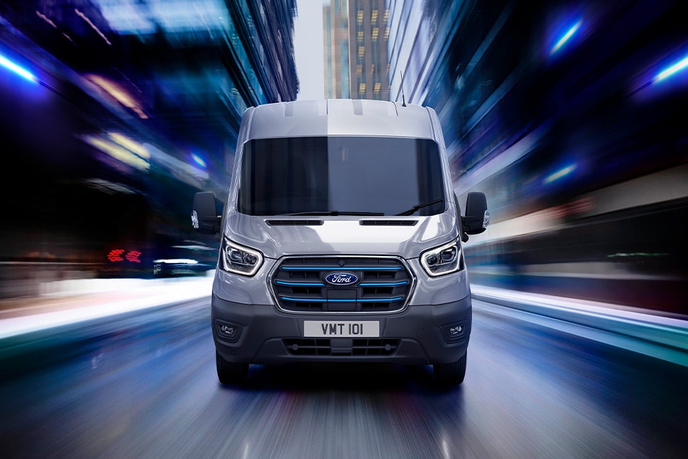 Een grijze Ford Transit-bestelwagen met blauwe grille-accenten rijdt 's nachts met hoge snelheid door een stad.