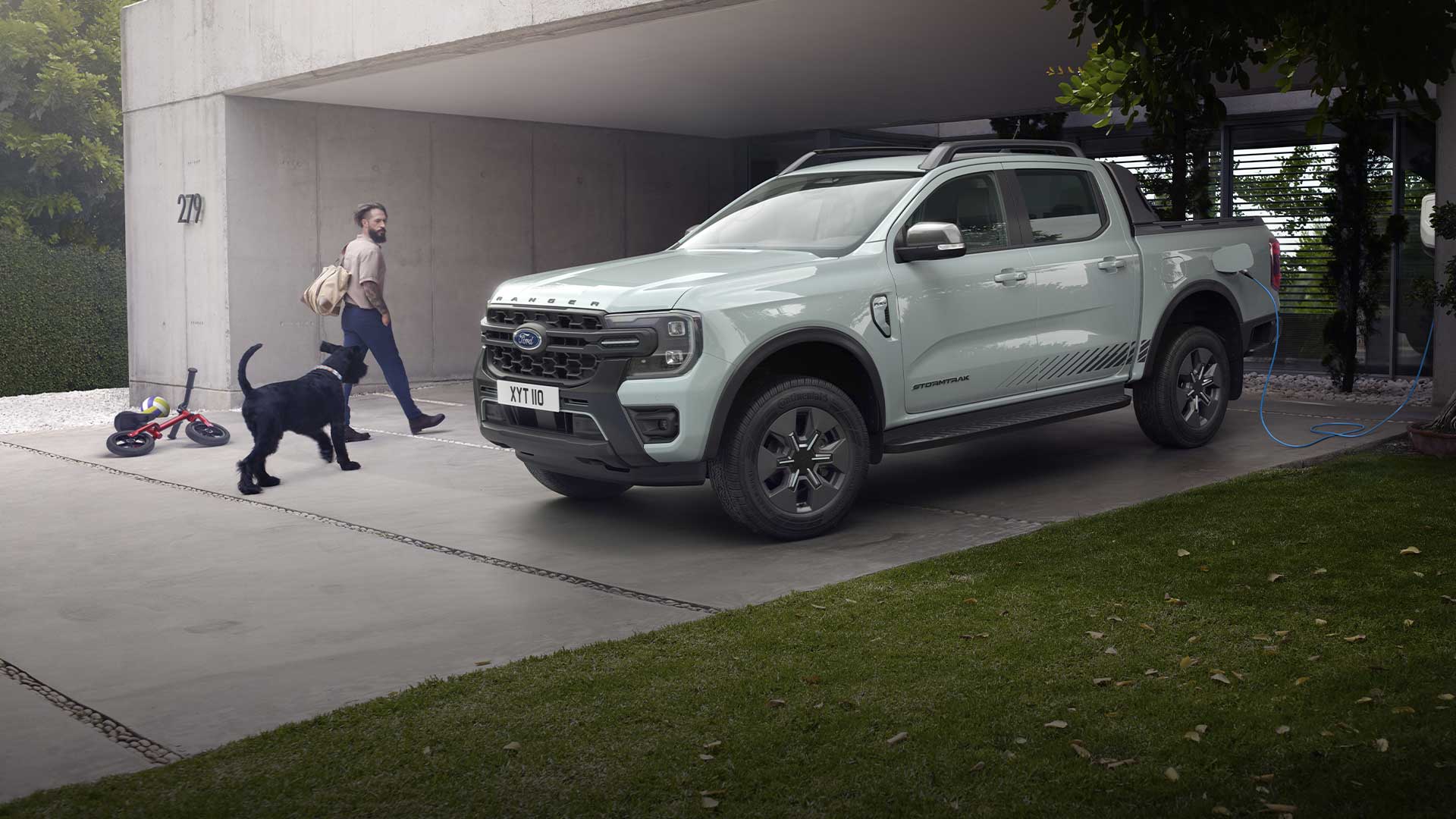 Een man en een hond lopen naar een grijze Ford Ranger Plug-in Hybride pick-uptruck die op een oprit opgeladen wordt. 