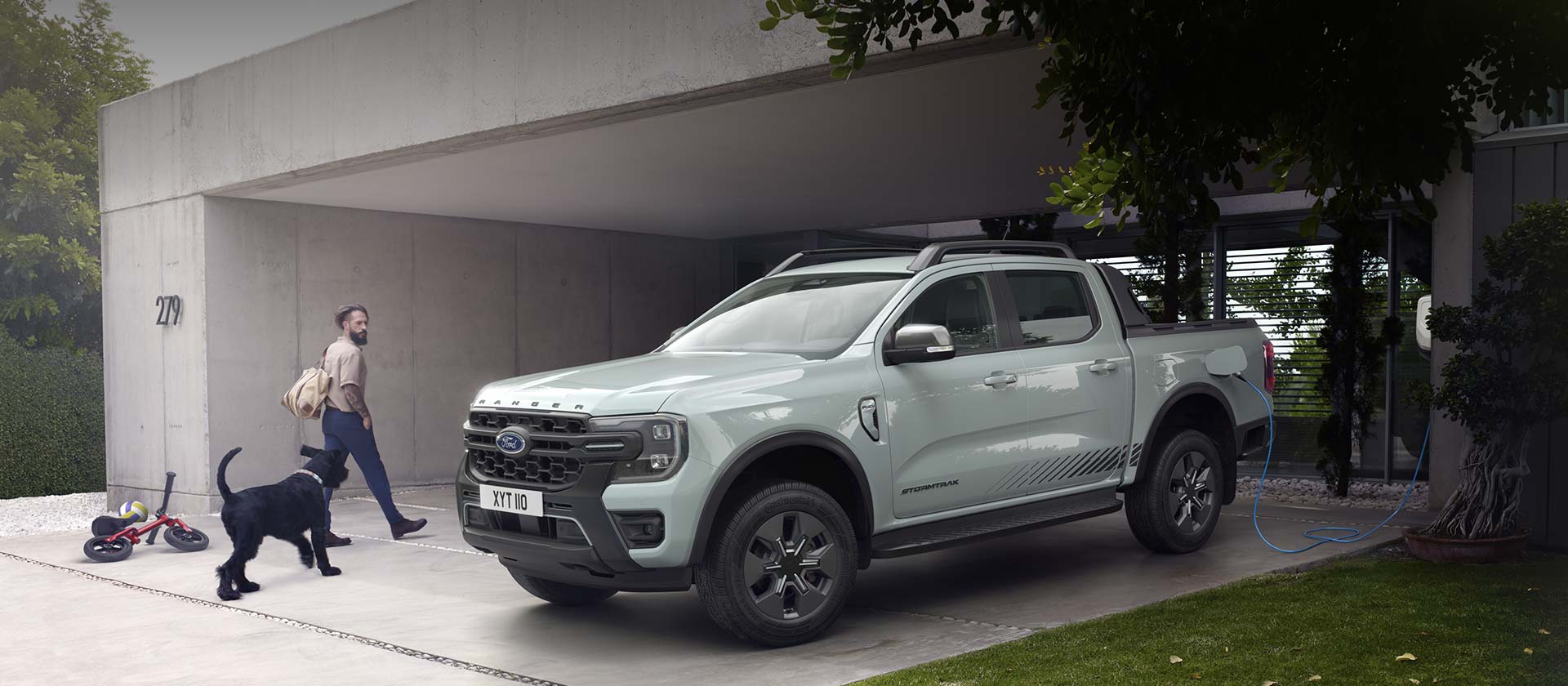 Een man en zijn hond lopen een modern huis binnen en passeren een Ford Ranger Stormtrak PHEV die wordt opgeladen. 