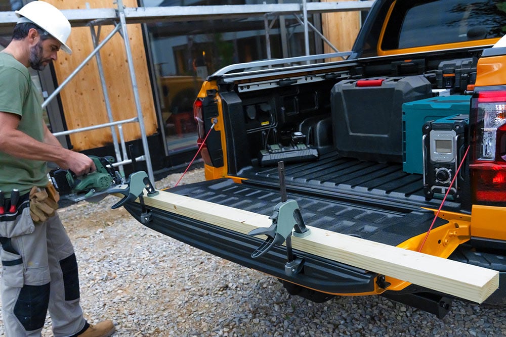 De achterklep van een gele Ford Ranger is open, met een houten plank die er met een klem aan vastzit.