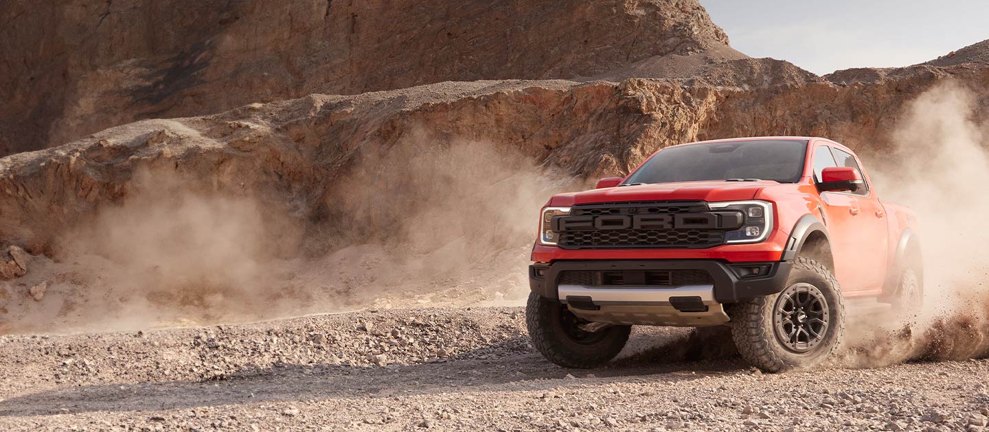 Een rode Ford Ranger Raptor laat een stofwolk achter terwijl hij door een woestijnlandschap rijdt.
