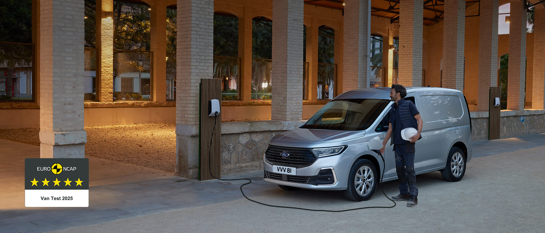 A man holding a hard hat plugs a wall box charging cable into All-New Ford Transit Connect in Stardust Silver. 