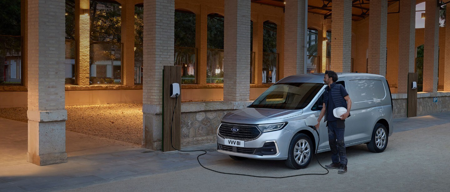 Een man met een helm steekt een laadkabel van een wallbox in de nieuwe Ford Transit Connect in Stardust Silver.