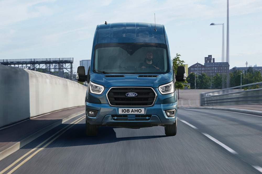 Vooraanzicht van een blauwe Ford Transit-bestelwagen die op een weg rijdt.