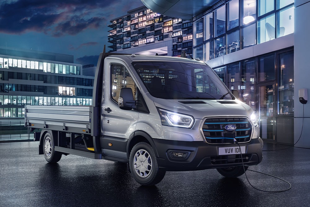 Een zilverkleurige Ford E-Transit-chassis met cabine die 's nachts geparkeerd staat voor een modern gebouw.