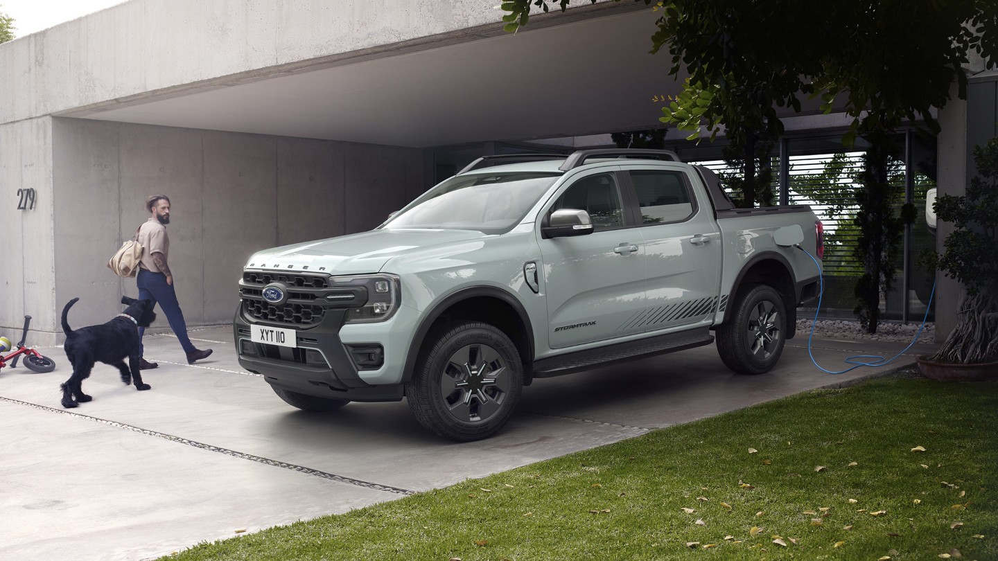 Man en hond lopen naar een modern huis langs een geparkeerde Ford Ranger Stormtrak plug-in hybride die aan het opladen is.