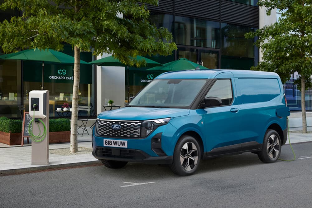 Voorzijde in driekwartaanzicht van een blauwe Ford E-Transit Courier, geparkeerd naast een laadstation.
