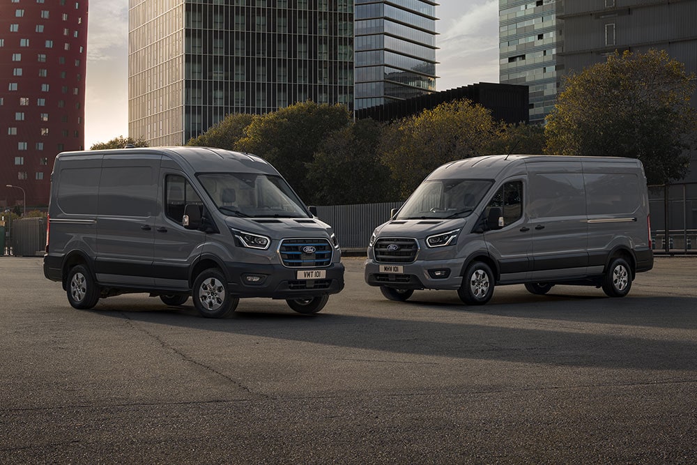 Twee Ford Pro Transit bestelwagens, waarvan één elektrisch, geparkeerd op een terrein met moderne gebouwen op de achtergrond.