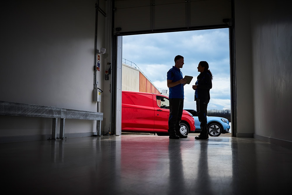 Twee mensen staan te kletsen bij een garagedeur, met een rode Ford-bestelwagen en een blauwe Ford SUV die buiten geparkeerd staan.