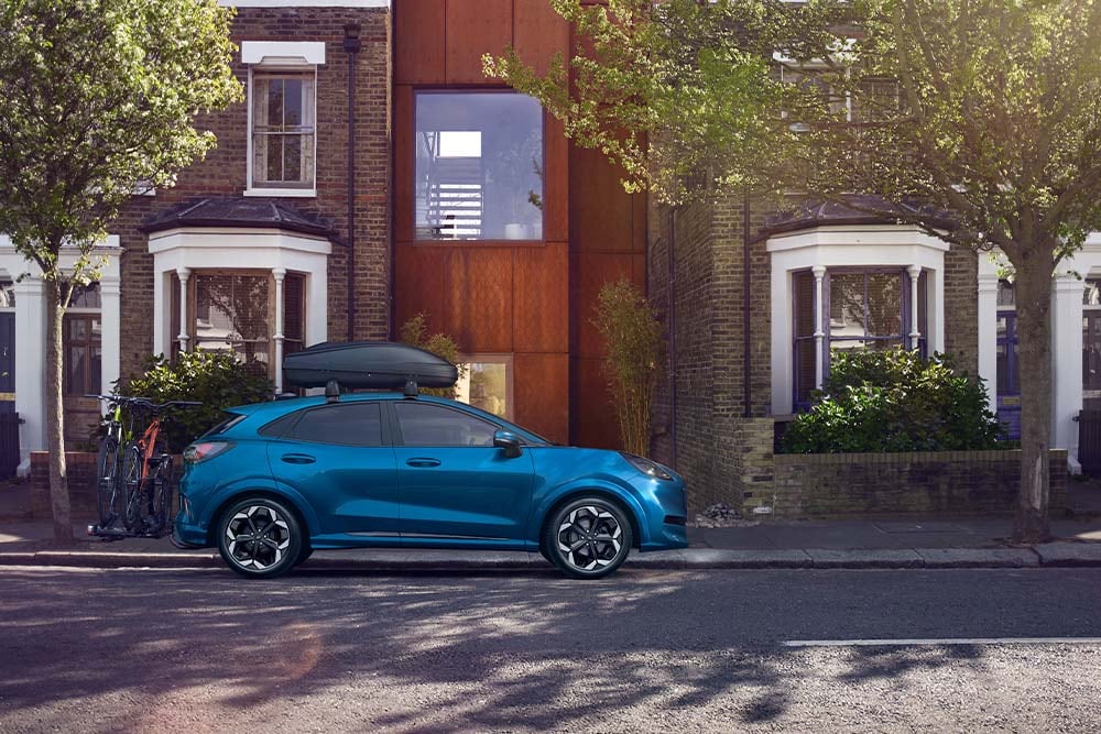 Zijaanzicht van een blauwe Ford Puma met een imperiaal en fietsendrager, geparkeerd in een woonwijk.