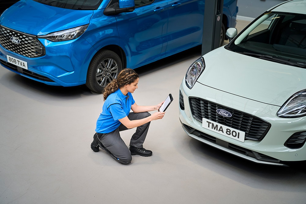 Een techneut zit tussen een blauwe Ford-bestelwagen en een witte Ford Puma en kijkt naar een tablet.
