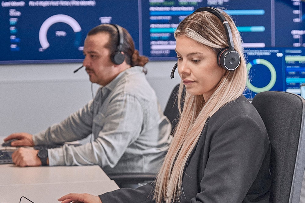 Twee personen met headsets gericht op computerschermen in een moderne controlekamer, met datadisplays.